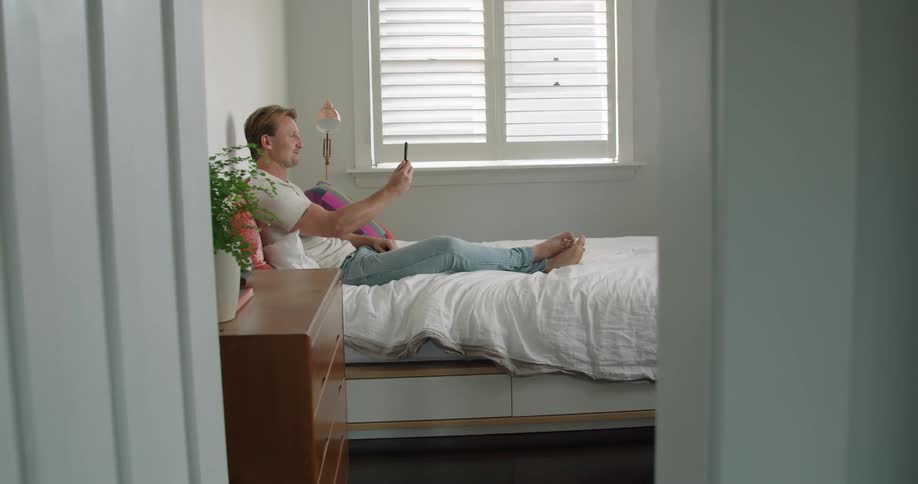 Man greets someone on a video call on his smartphone while resting in bed at home