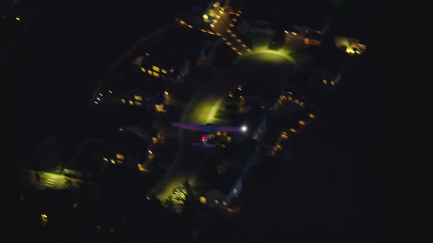 Rare top down high altitude night aerial shot of a white single engine airplane flying over mixed rural and residential landscape with anti collision strobe lights flashing