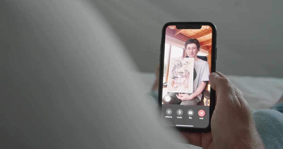 A man resting on his bed is shown an artwork by another man through video chat on his smartphone