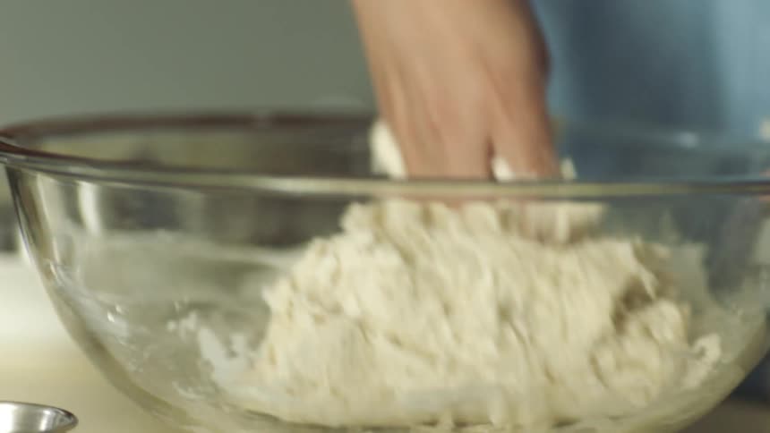 A person in a room kneading dough in a mixing bowl on a table