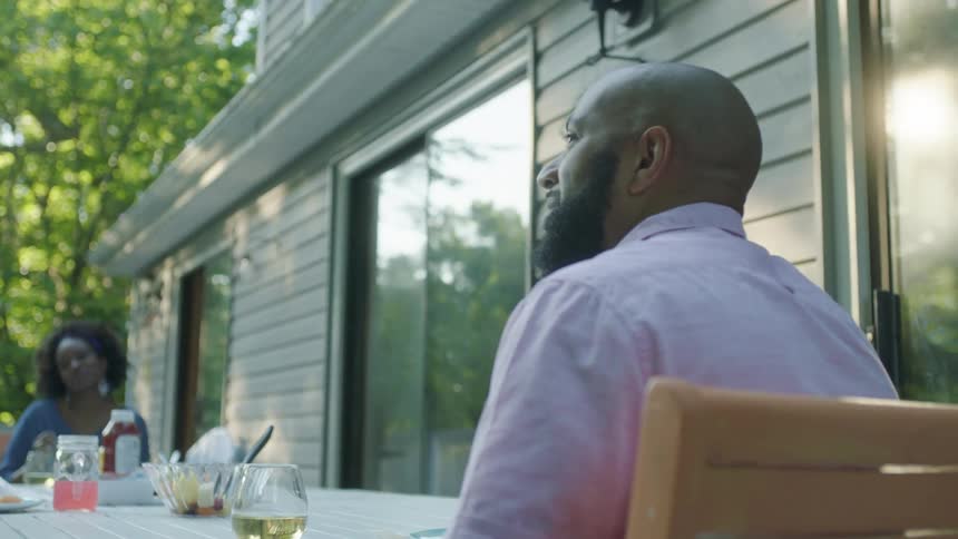A man sits outside at a table in the backyard of a house with people eating and smiles