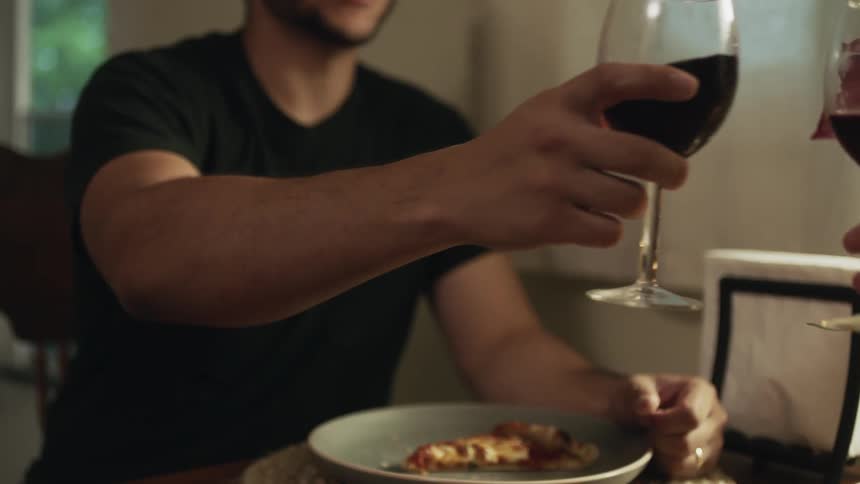 A man picking up a piece of pizza and celebrating with wine at a dinner table
