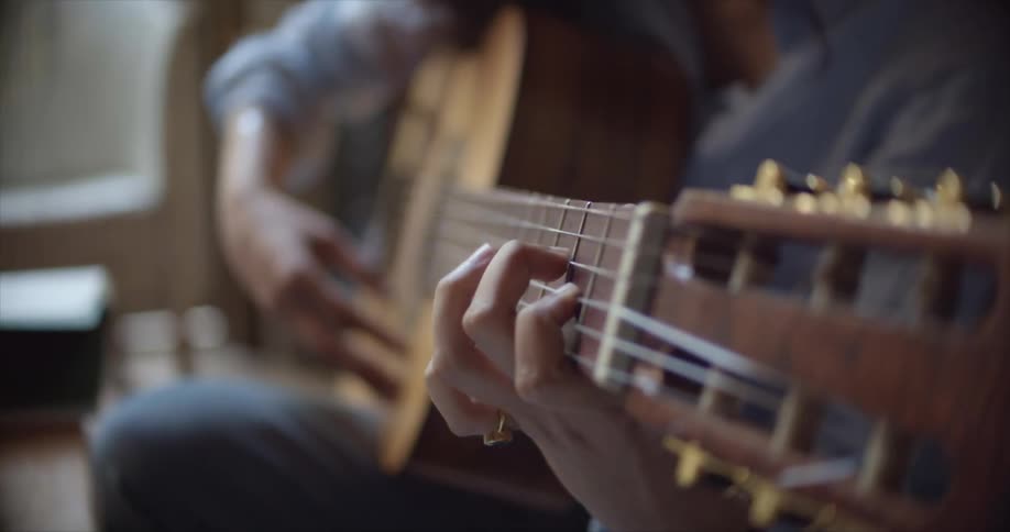 A woman in a room sitting and playing a guitar with a couch in the background