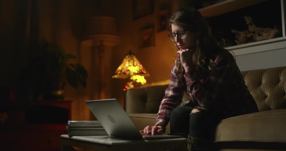 A woman types on a laptop computer while sitting on a couch in a living room at nighttime