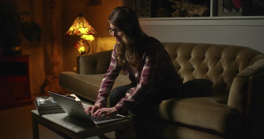 A woman types on a laptop resting on a coffee table while sitting on a sofa in a dimly lit living room at night