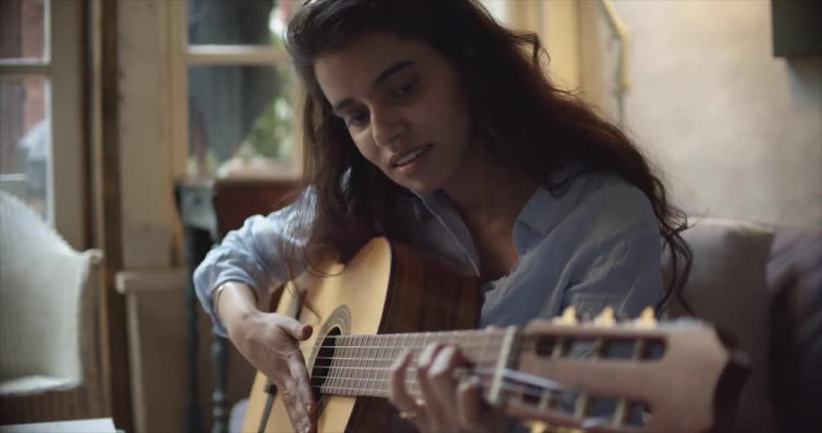 A young woman playing an acoustic guitar while sitting on a living room sofa