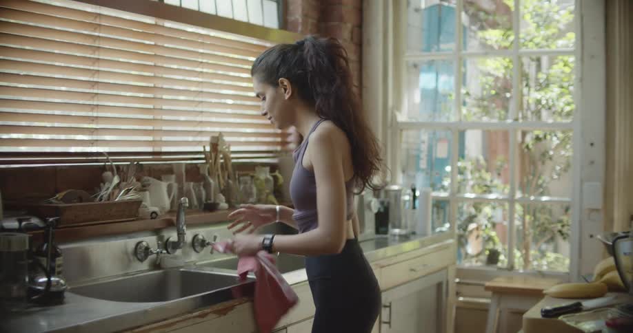 A woman wearing workout gear washes and dries her hands at the kitchen sink
