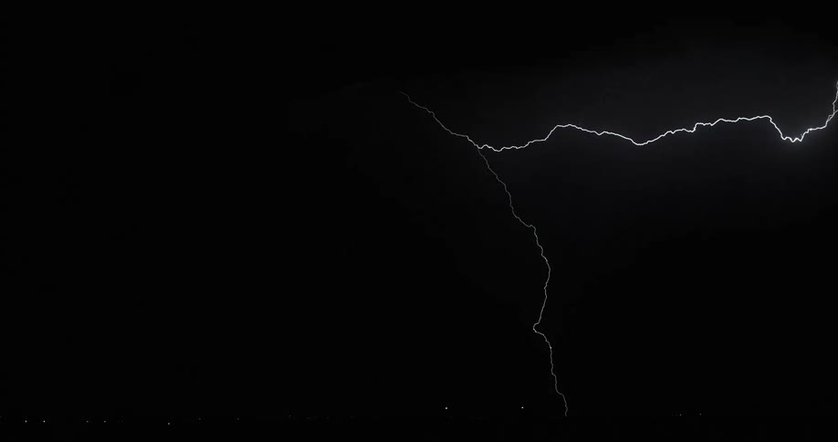 Lightning forks across the sky in storm clouds and strikes down to the ground at nighttime