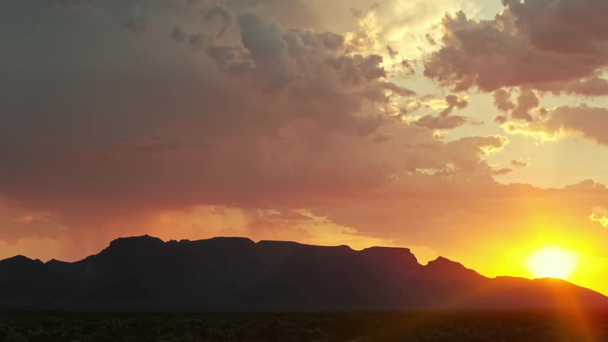 Lightning flashes in clouds in the sky over a sun setting in the distance behind mountains