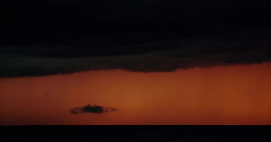 Lightning strikes down from the center of a dark storm cloud in an orange sky at sunset