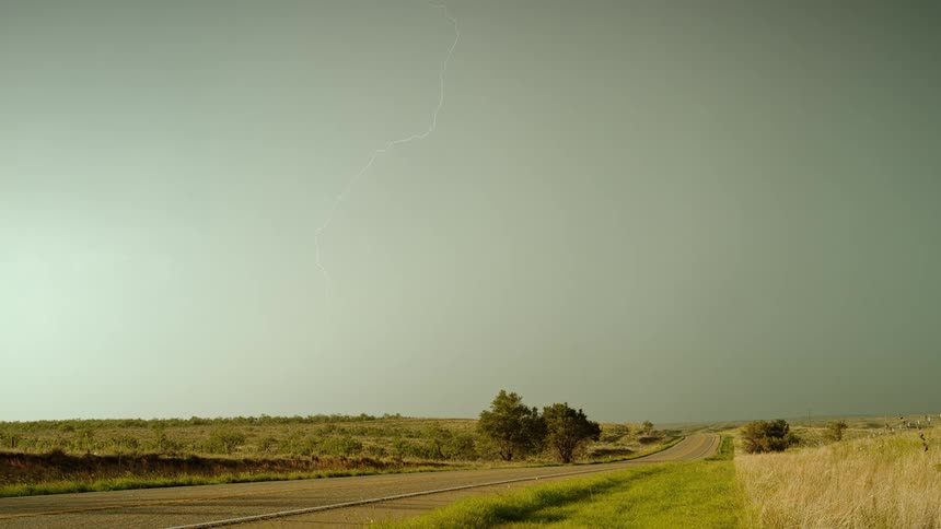A road next to a grassy plain with trees lightning the sky and clouds in the background