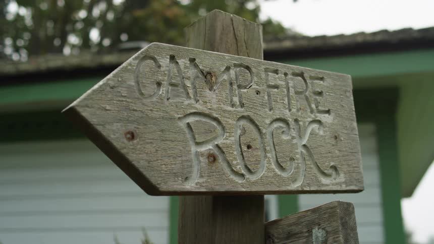 A signed carved in wood with a building trees the sky and clouds in the background