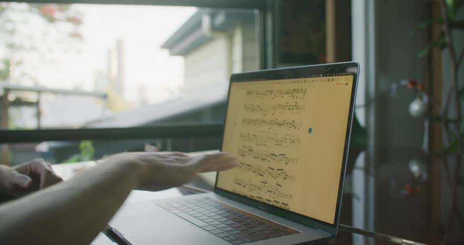 A man stands next to a man sitting at a grand piano as they sing a song piece displayed in front of them on a laptop indoors at daytime
