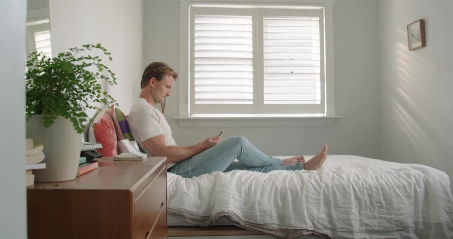 A man in a bedroom sitting on a bed looking at a smart phone with a wall and window in the background