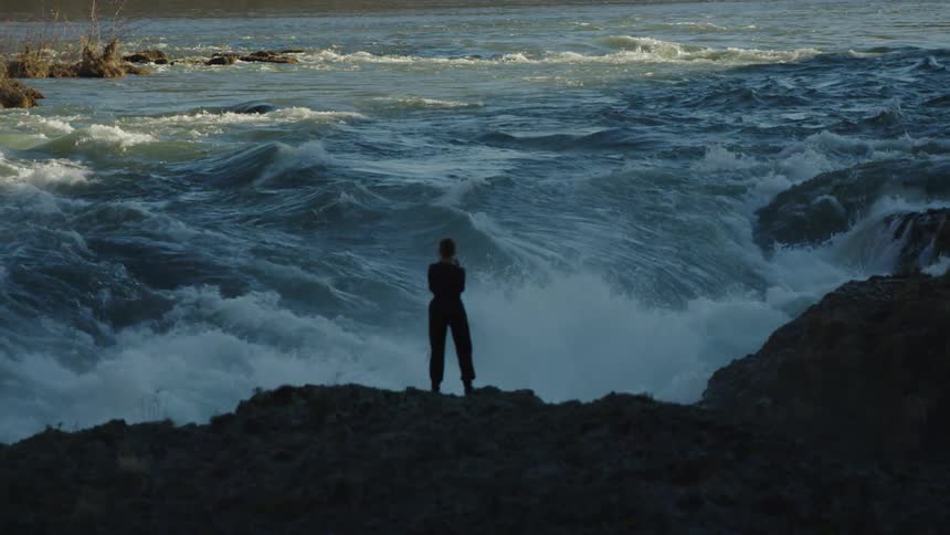 A woman stands at the edge of a small cliff watching river rapids pour down into a waterfall silhouetted against the river whitewater rapids