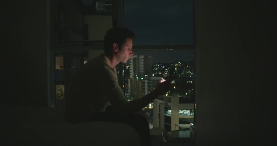 A man sits in his home and scrolls on his cell phone and looks out at the skyline