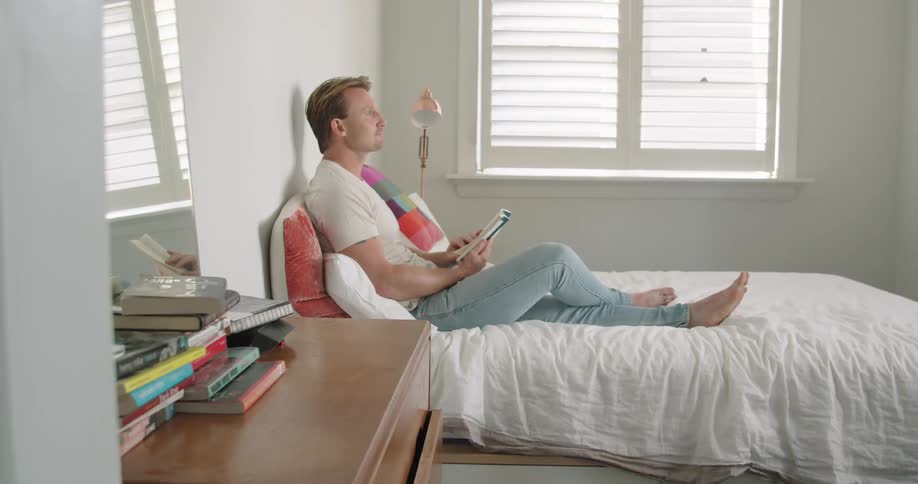 Man reading a book over the bed in his bedroom at home
