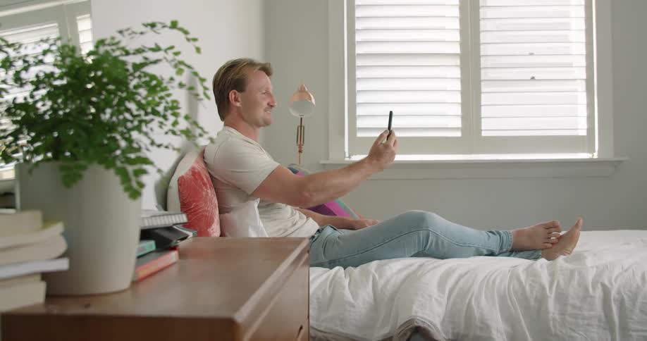 A man in pajamas smiling while video chatting on a smartphone in bed