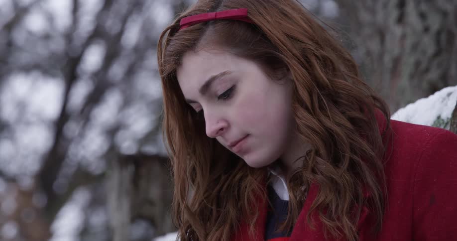 A red haired woman in a red coat sits in a snow-covered landscape looking at something below camera