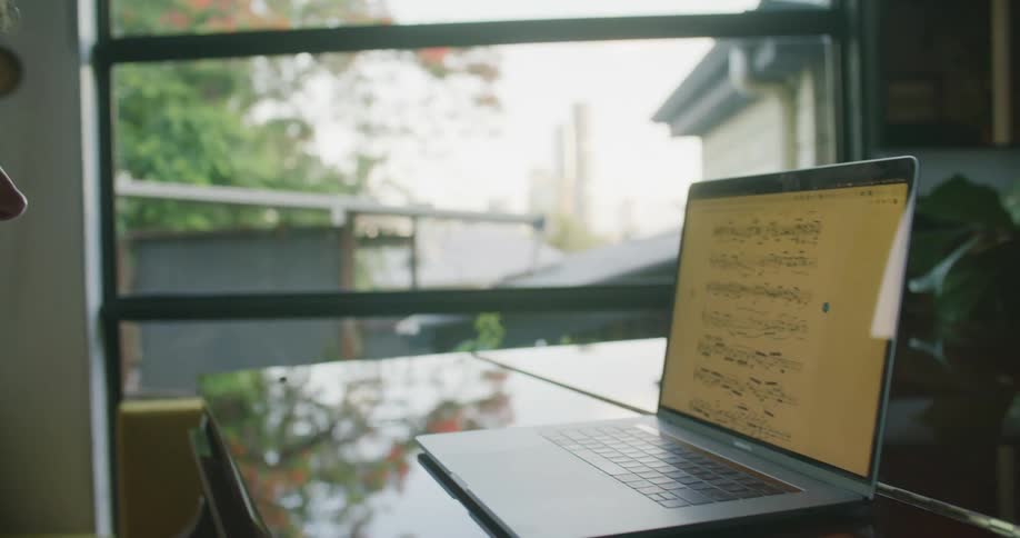 A white male plays a piano while reading sheet music displayed on a laptop