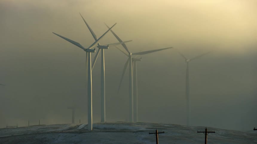 Windmills rising from the fog rotate as the camera pushes down a rural road with power poles visible in the bottom right side of the frame at sunrise in rural clean energy America