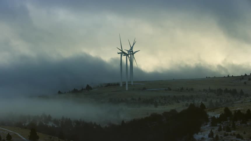 Low valley fog and thick storm clouds sandwich these wind turbines at a wind farm in a picturesque and dynamic clean energy scene