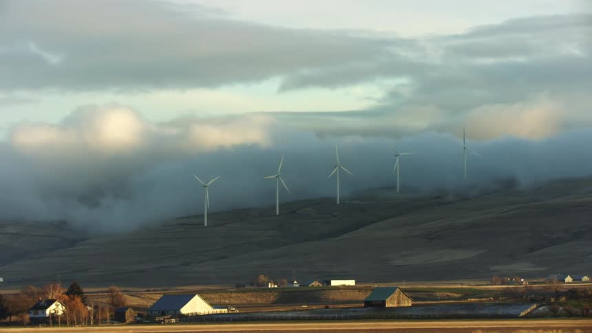 Five wind turbines rotate on a ridge above a pastoral rural farm valley at sunrise on a cold winter morning with low valley fog