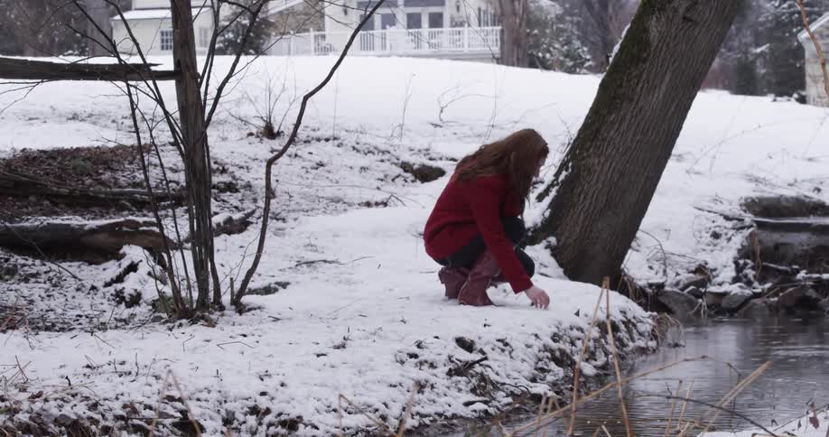 A woman crouches down in the snow by a stream