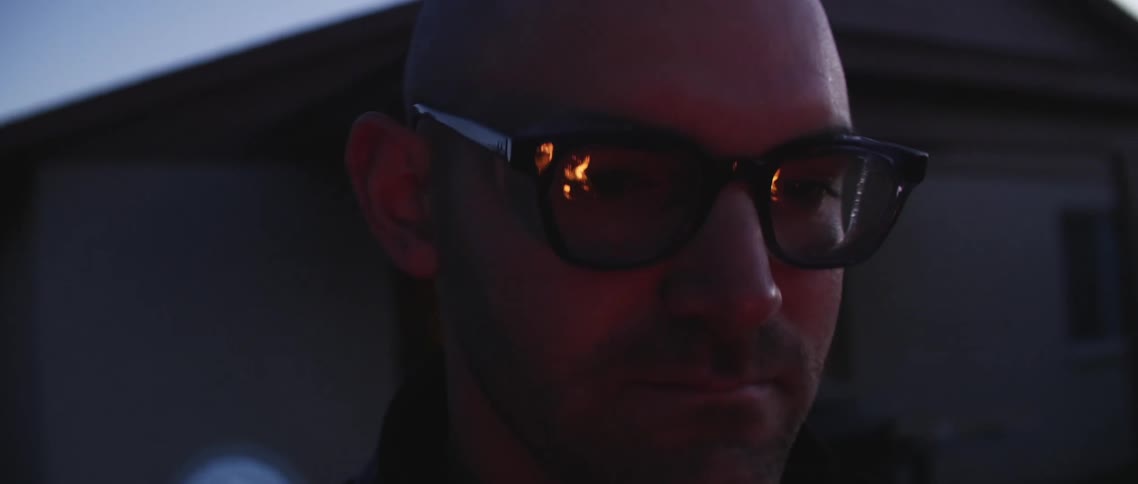 A man with glasses with fire reflecting off of it with a house the sky and clouds in the background