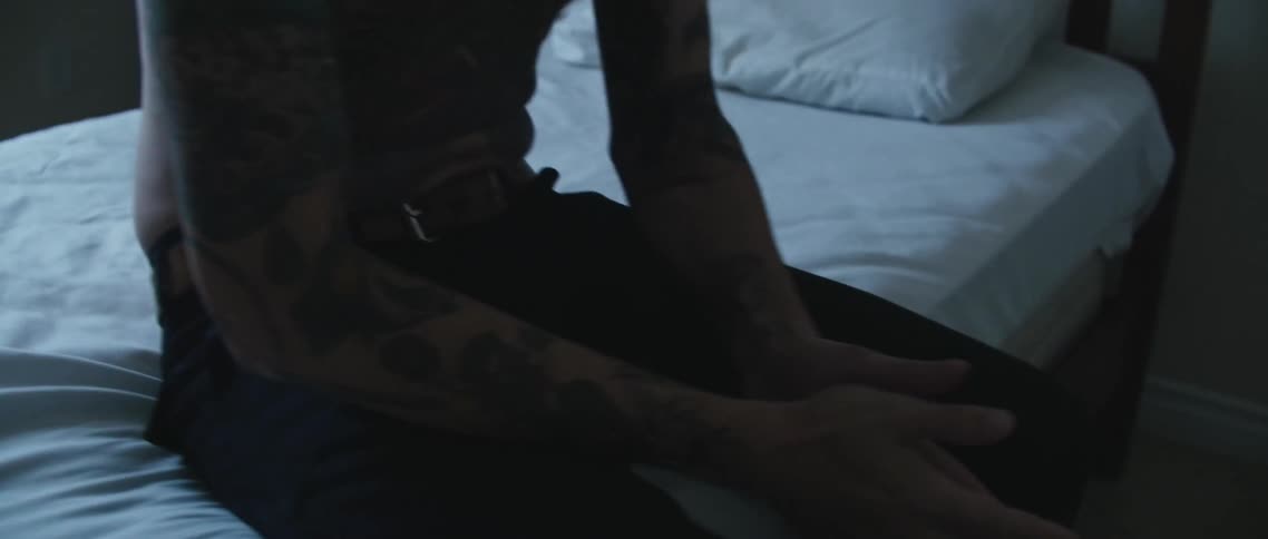 Shirtless bald man with tattoo sits down on bed in the bedroom