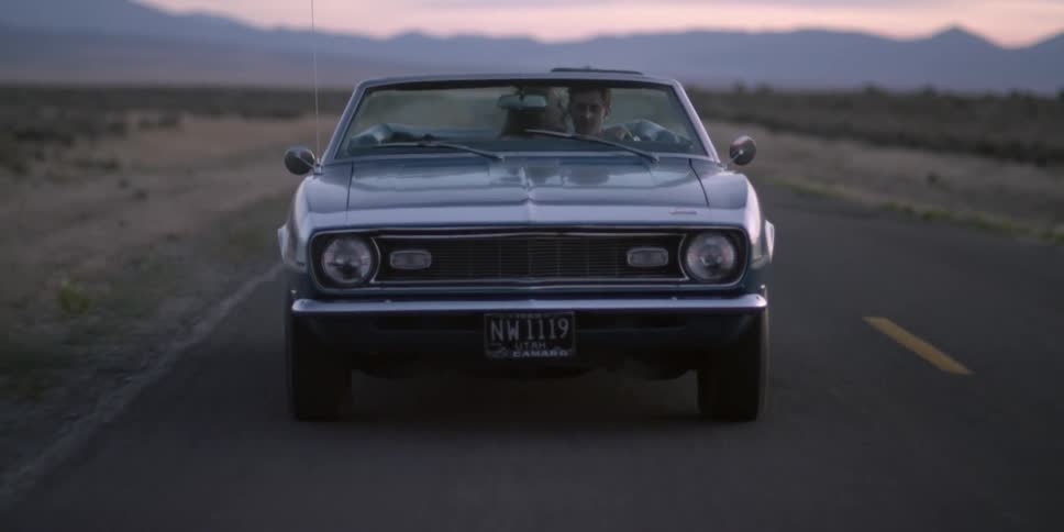 A woman kissing a man while he drives a convertible with the sky and clouds in the background