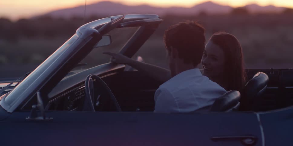 A man and a woman in a convertible with the landscape the sky and clouds in the background
