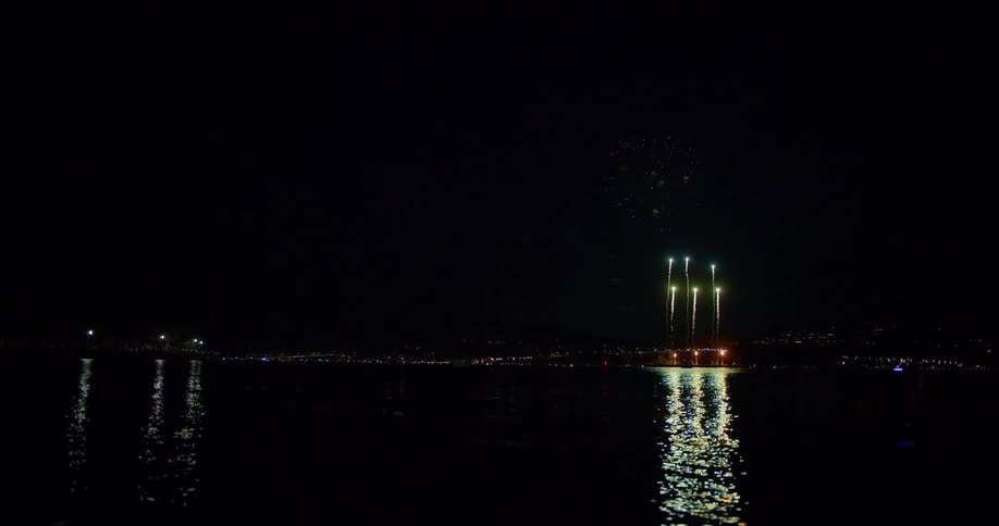 Different color fireworks go off in the distance of a body of water over a city skyline at nighttime