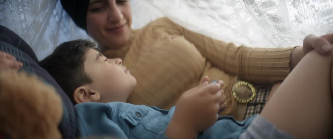 A mother and son sit on the ground with pillows under a white blanket made into a tent as the boy holds something in his hands