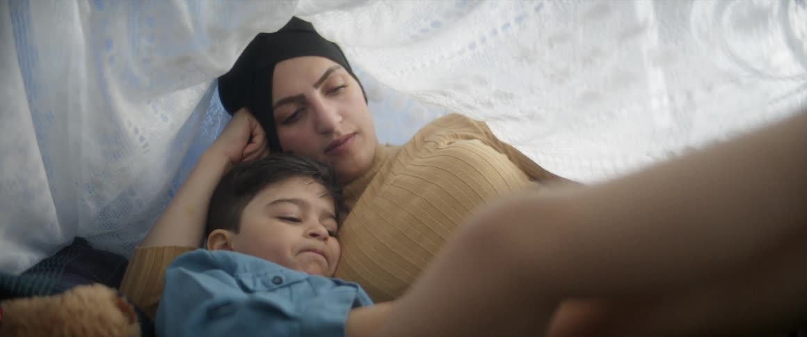 A woman lies down with her son and strokes his hair as he plays with a cell phone