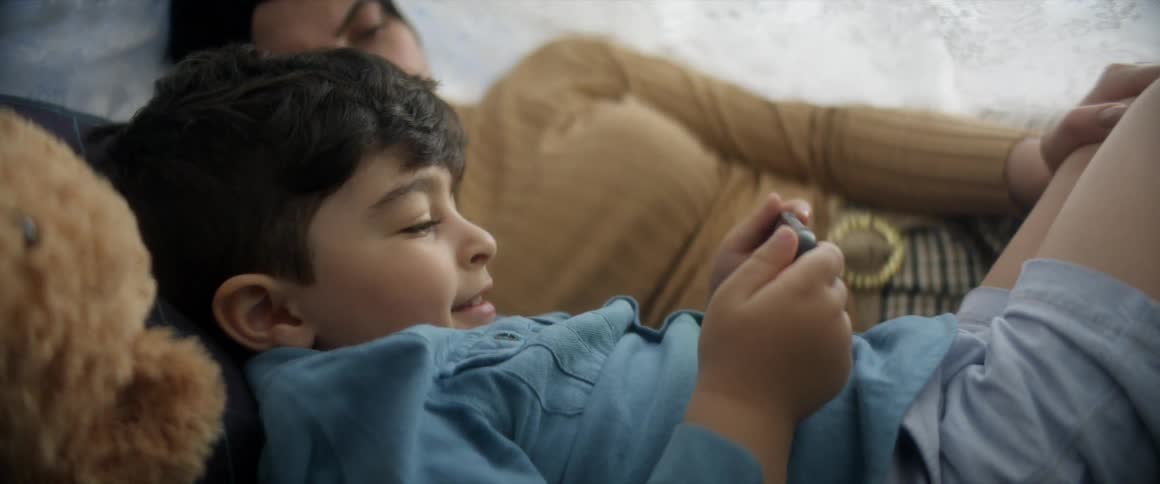 Mother and son are laying in a fort outdoor in the breeze and playing on cell phone