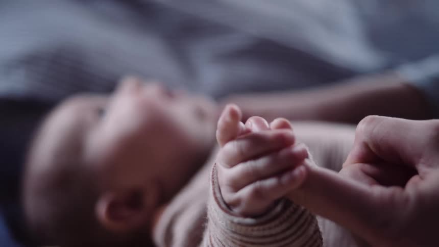 A baby lying on a bed plays with the finger of an adult