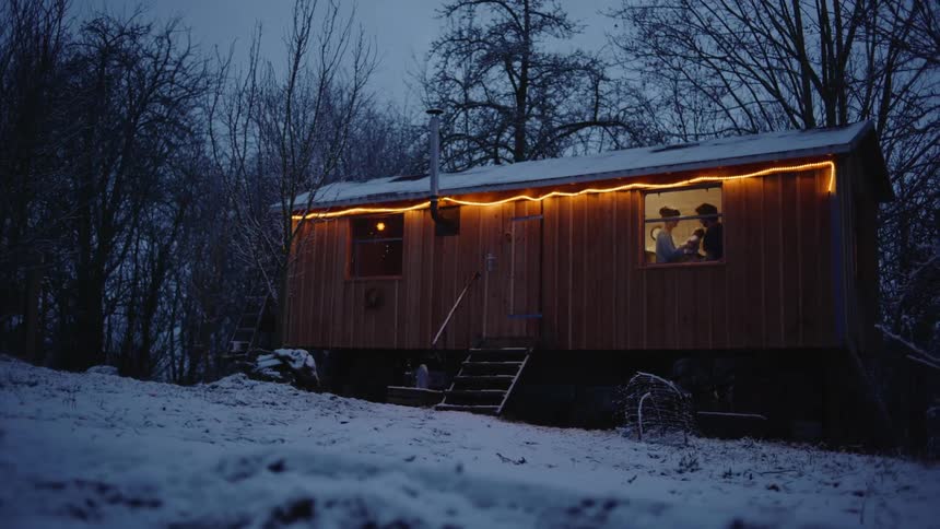 A woman in a tiny house holds a baby up in the air and a man leans forward and kisses the woman
