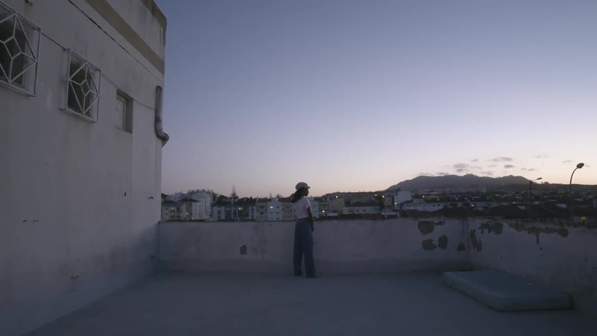 A woman with a hat standing on a rooftop with buildings the sky and clouds in the background