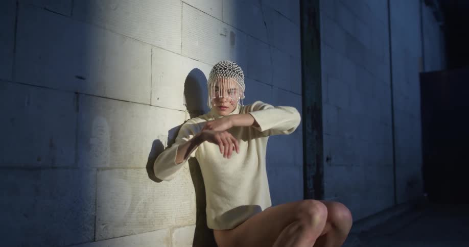 A woman wearing a leotard and beaded veil dances against a wall in a sexy manner