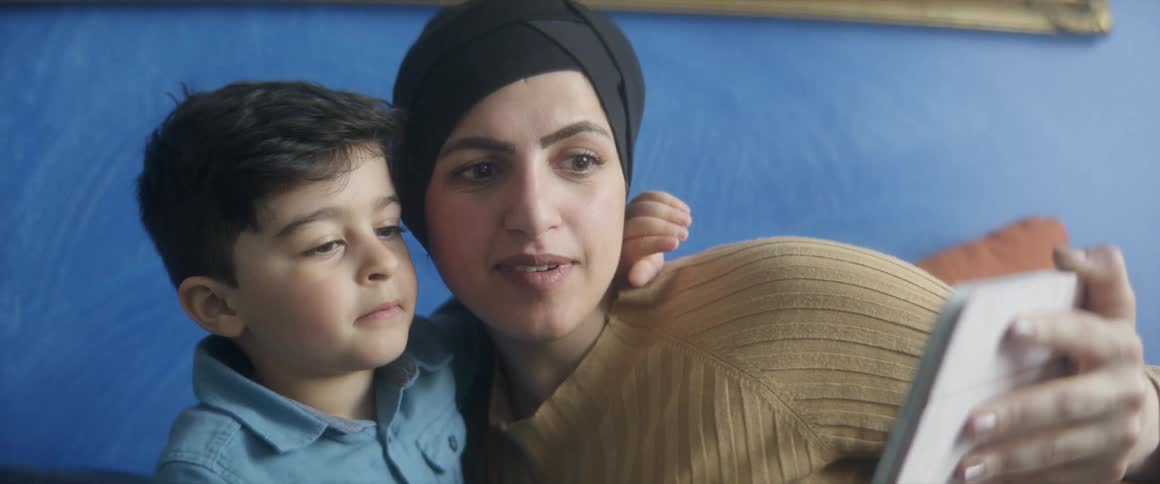 A woman smiles and poses with her son as she films them with her tablet