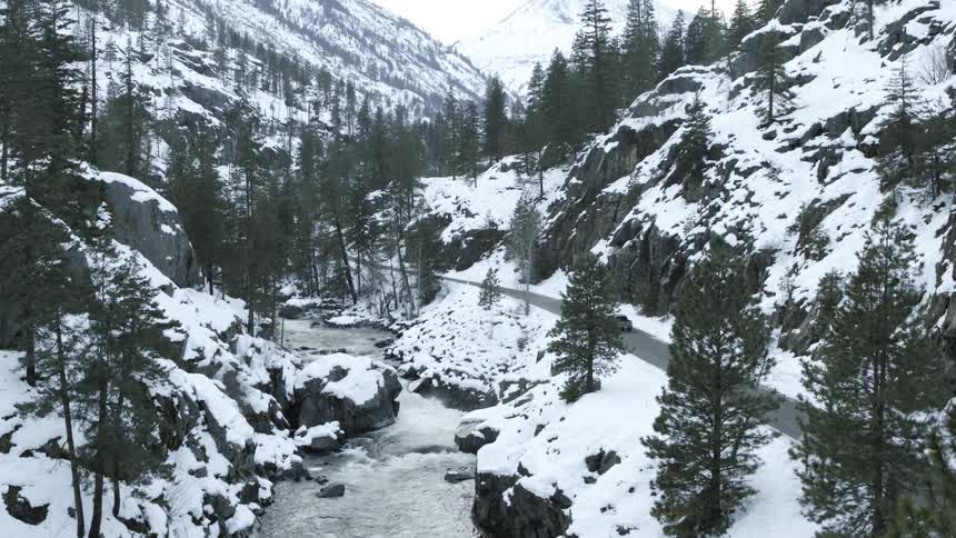 A pickup truck drives on a forest road next to a rushing river and snow covered mountains ahead