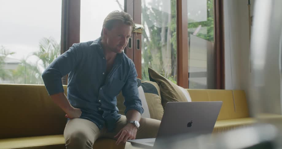 A man participates in a video chat using a laptop while sitting on a sofa in the living room of a house during the day