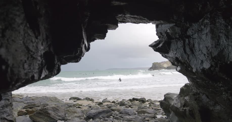 Ocean waves onto the shore outside of a stone cave on a beach covered in rocks