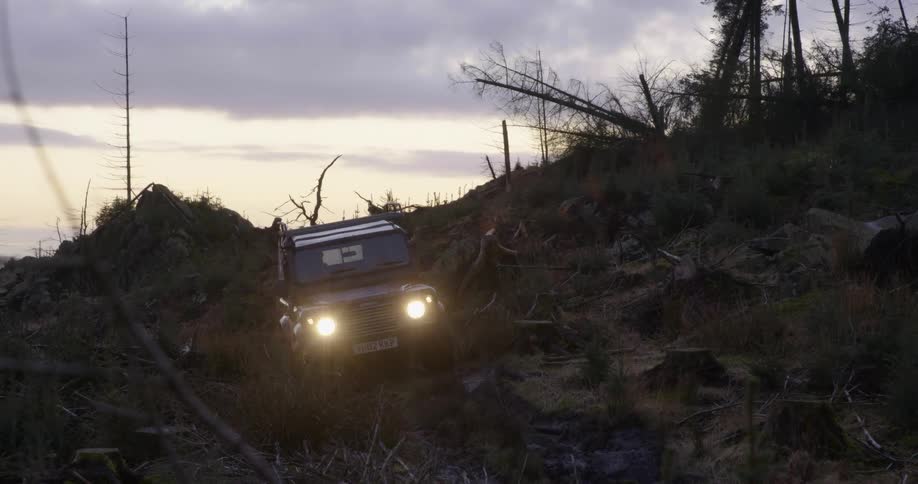 A land rover with headlights on makes its way downs a slope over rocks and tree stumps