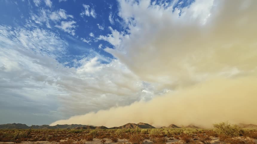 Clouds roll in overhead in a desert while a sand storm follows behind and overtakes the sky and the ground below