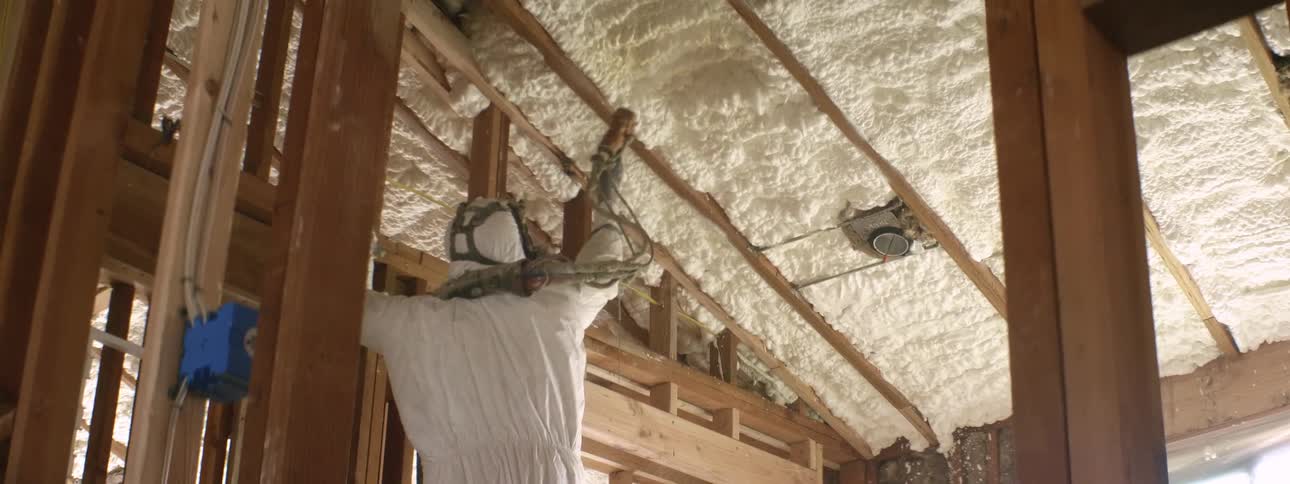 A worker wearing protective gear applies insulating foam in the roof beams of a house