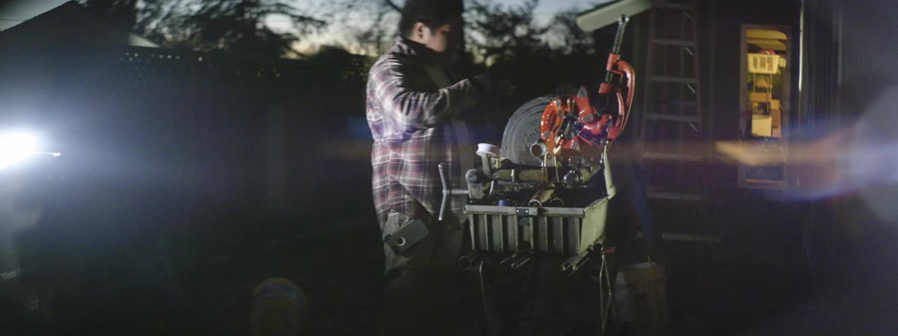 A man oils a removed part of a machine before replacing it while standing outdoors in the yard of a house at sunset