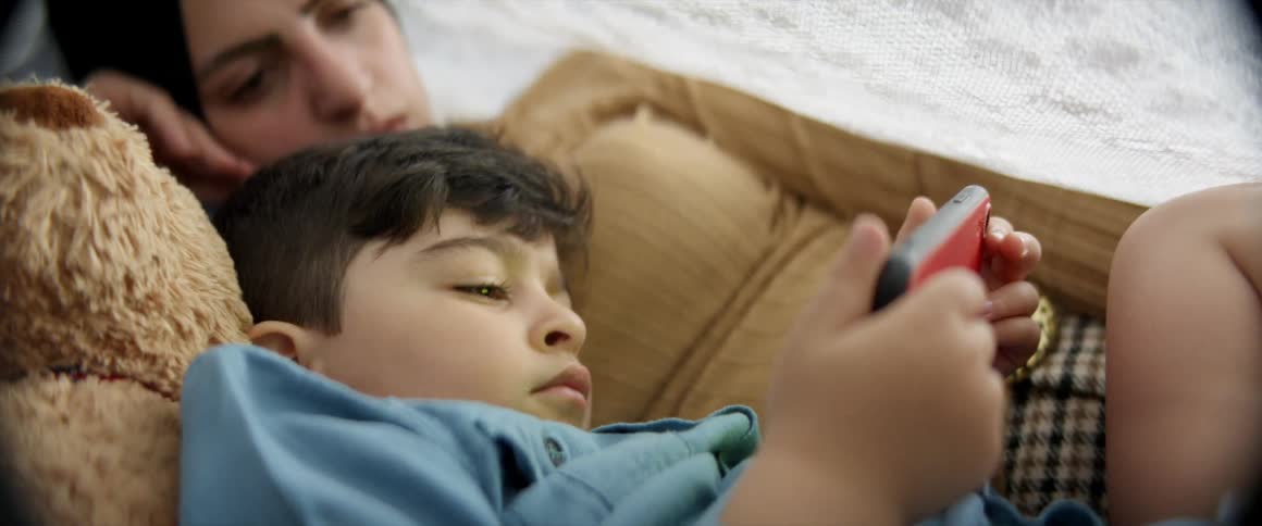 A woman lies down with her son as he plays a game on a cell phone