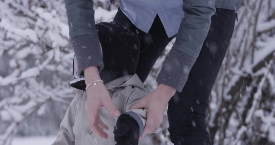 A mother secures a toddler's winter cap as they stand outdoors while snow is falling